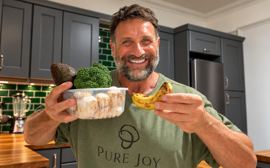PURE JOY co-founder Jordan smiling in his kitchen, holding mushrooms, avocado, broccoli and a banana.