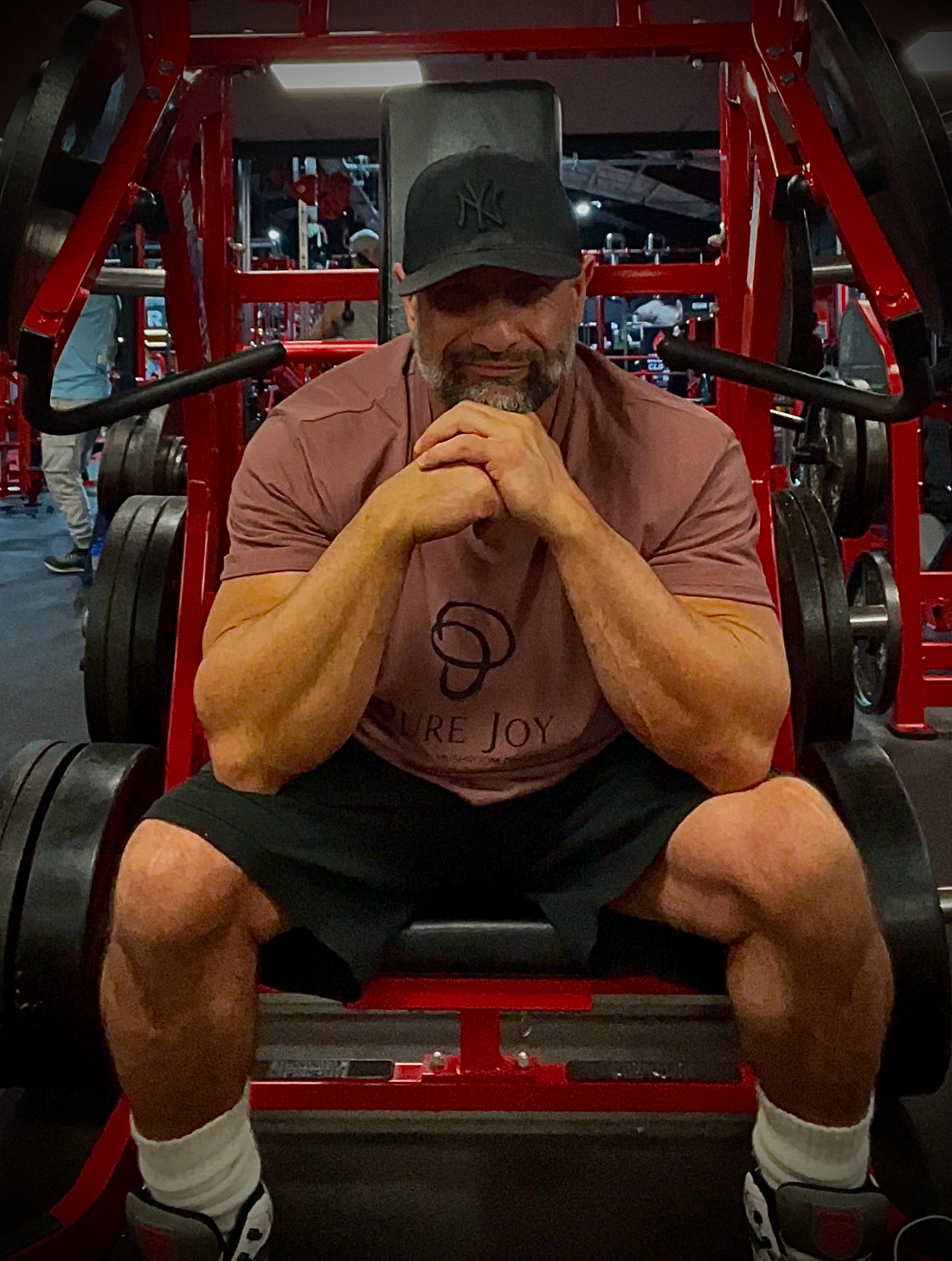 PURE JOY co-founder Jordan smiling while seated on gym equipment, wearing a PURE JOY T-shirt and black cap.