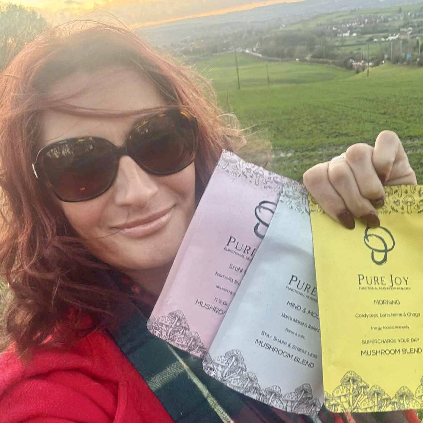 Customer holding Pure Joy Mushrooms Skin & Hair, Mind & Mood, and Morning Blend powders outdoors with a scenic landscape in the background.
